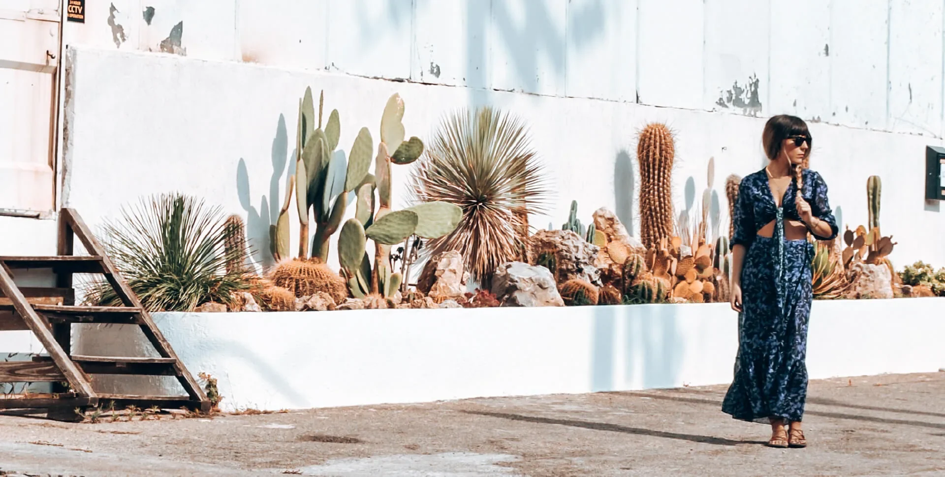 A woman in a long blue printed dress walking past a white wall lined with cacti