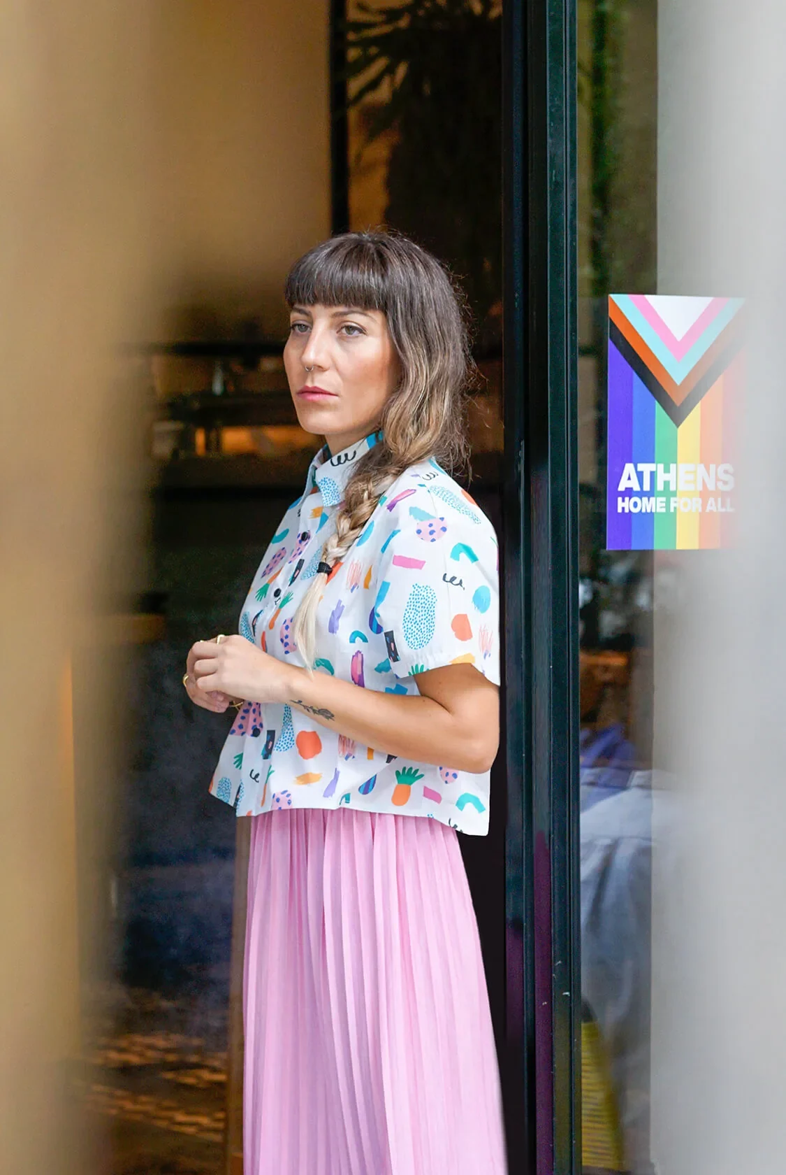 A woman in a cropped shirt with a painted-shapes print and a pink pleated skirt, leaning in a café doorway in Athens