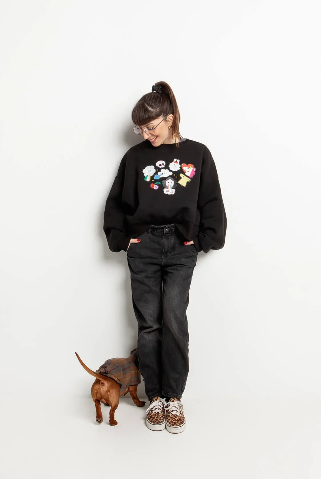 A woman in a black sweatshirt printed with a cluster of illustrated characters, black jeans and leopard sneakers, a small dog at her feet